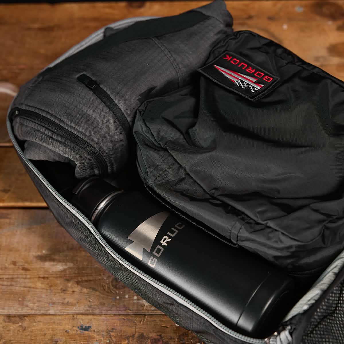 A Bullet Ruck Laptop - Ripstop ROBIC® - 16L from GORUCK sits on a wooden surface, showcasing a rolled-up jacket, folded shirt, and a branded metal water bottle. The partially zipped backpack provides a glimpse of its organized interior and ample lumbar support for enhanced comfort.