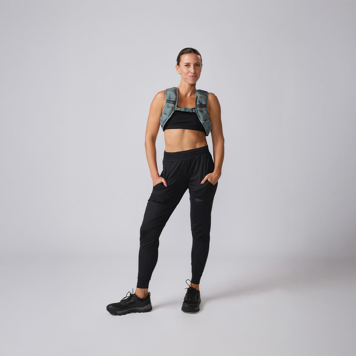 Woman in athletic wear and sneakers stands with hands in pockets, highlighting the Spy Ruck weighted vest—an essential piece for women's fitness gear—against a plain background.