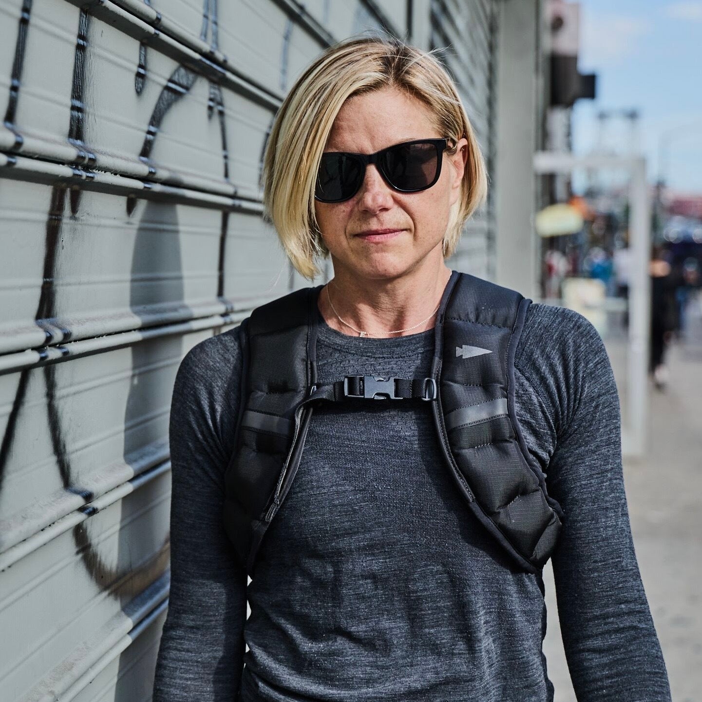 A woman with short blonde hair and sunglasses stands by a graffiti-covered metal gate, wearing the Spy Ruck black weighted vest—highlighting modern women’s fitness gear.