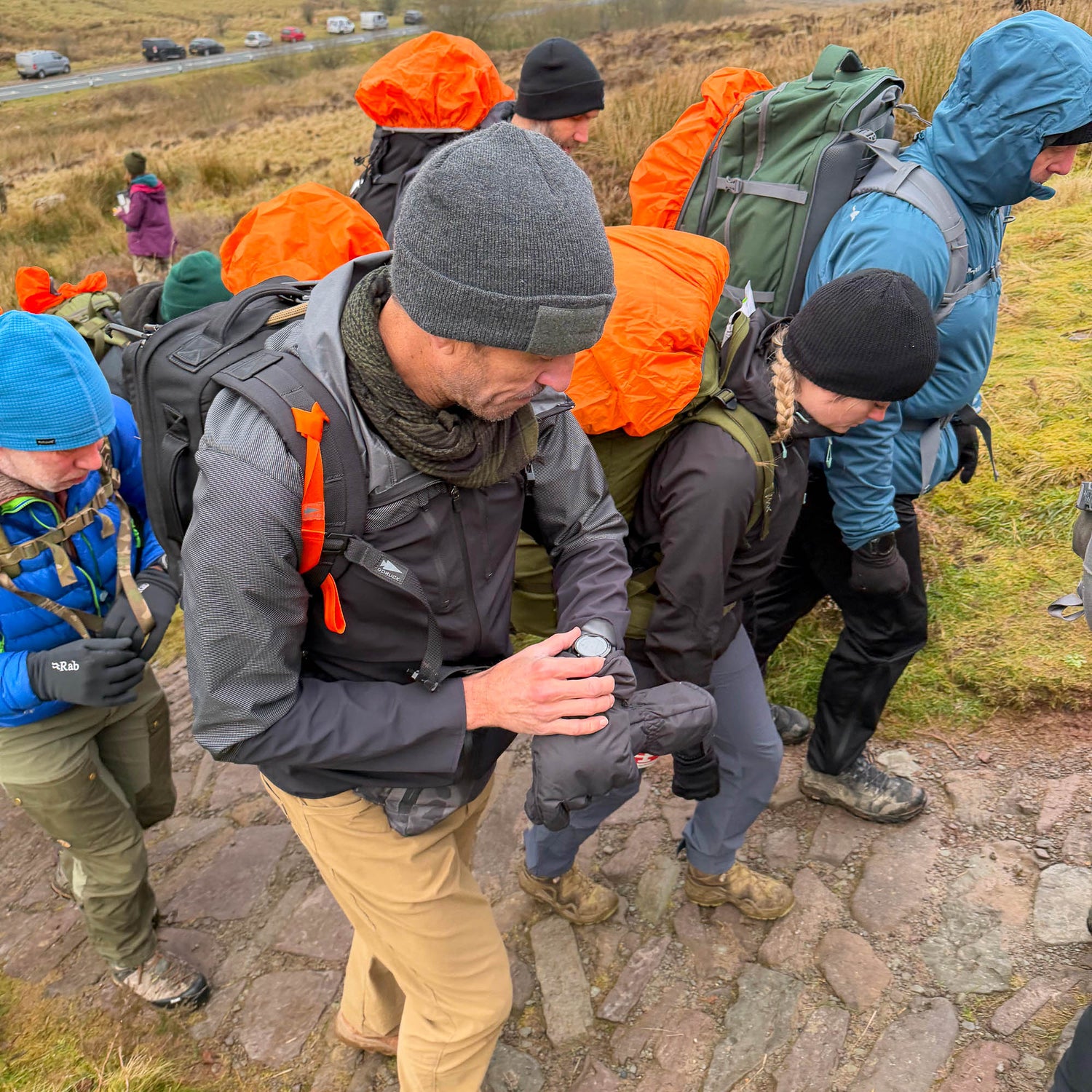 A group of hikers with backpacks and orange rain covers, wearing T-REX Hand Straps (pair) to reduce hand swelling, walk up a stone path on a grassy hillside.