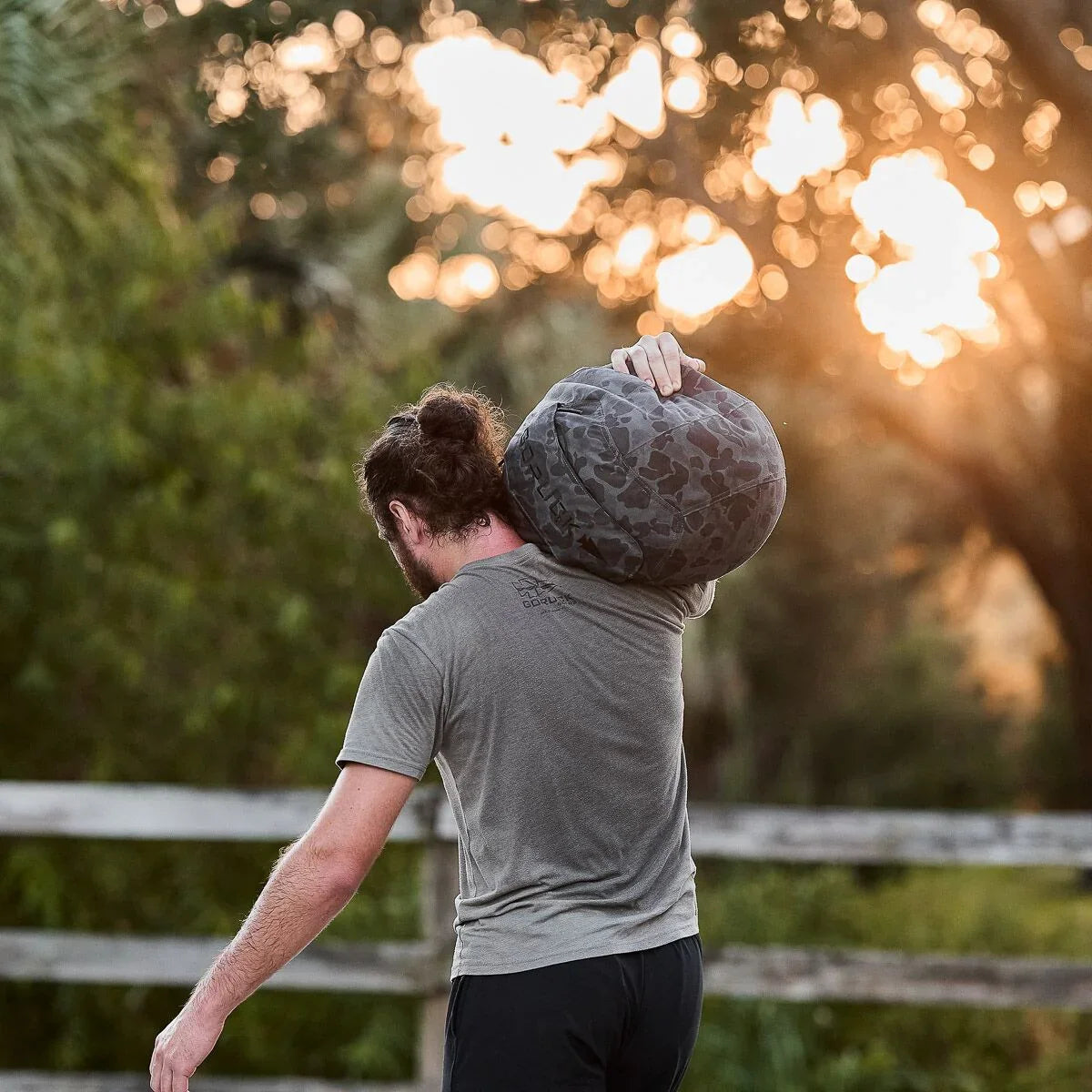 Man carrying a black camo GORUCK weighted bag on shoulder outdoors at sunset with wooden fence background