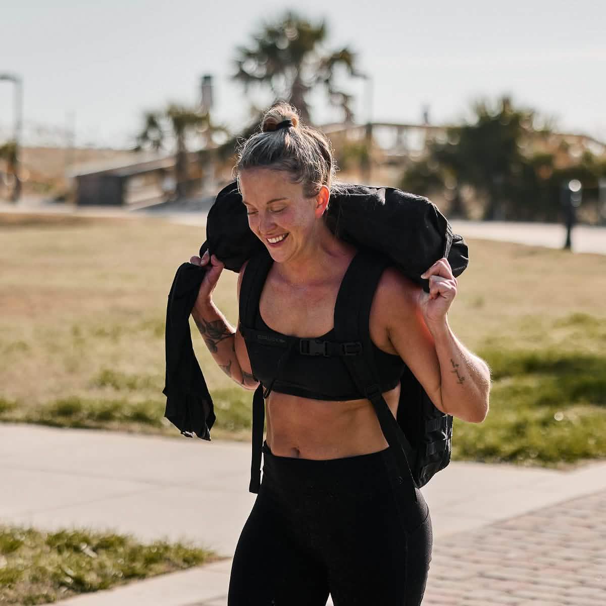 Athletic woman carrying GORUCK weighted sandbag outdoors on sunny day, training in black fitness gear