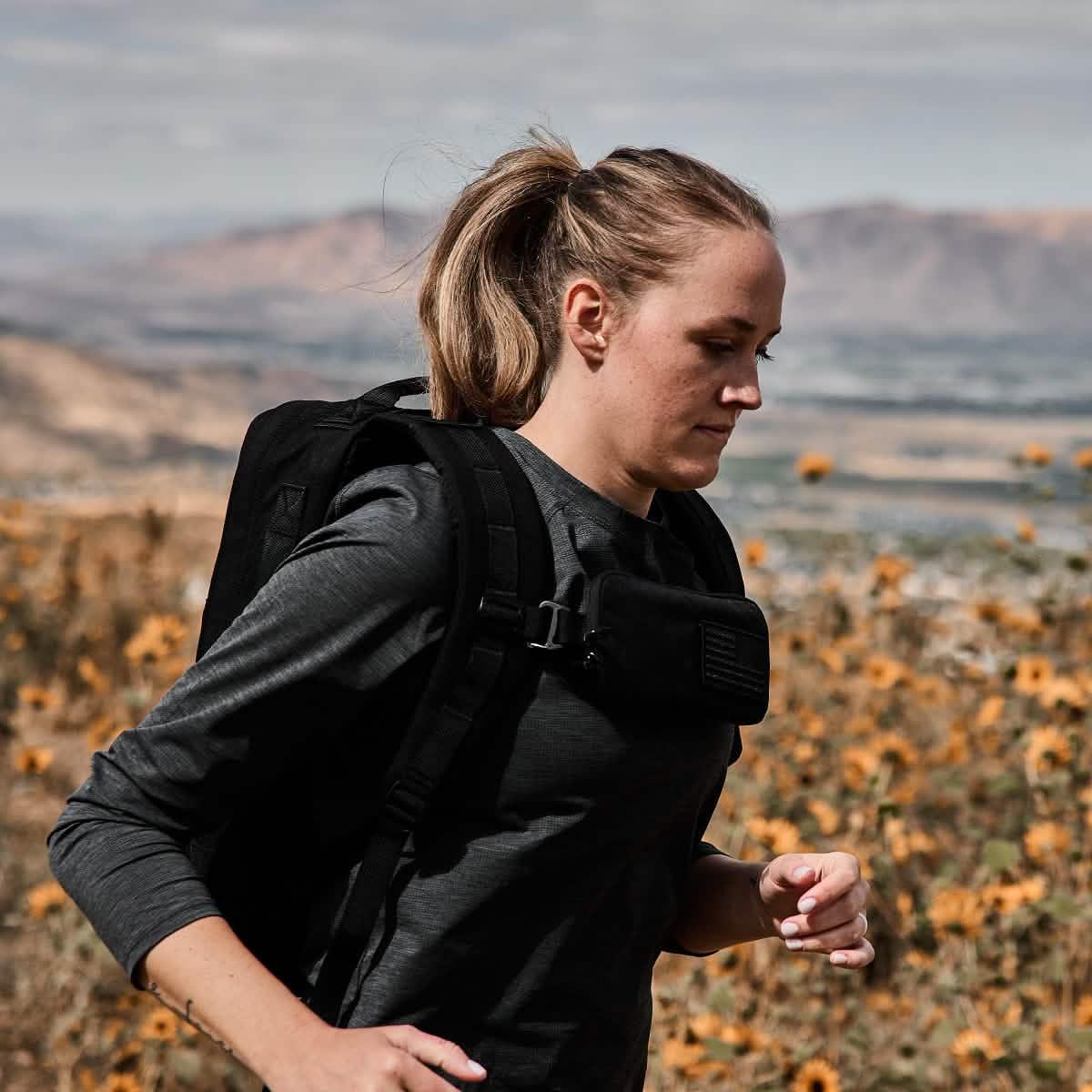 Woman rucking outdoors wearing a black sternum pouch and backpack, running in a field with mountains in the background