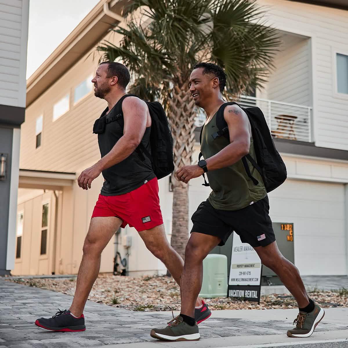 Two men carrying black rucksacks walking outdoors near modern homes with palm tree, wearing athletic gear