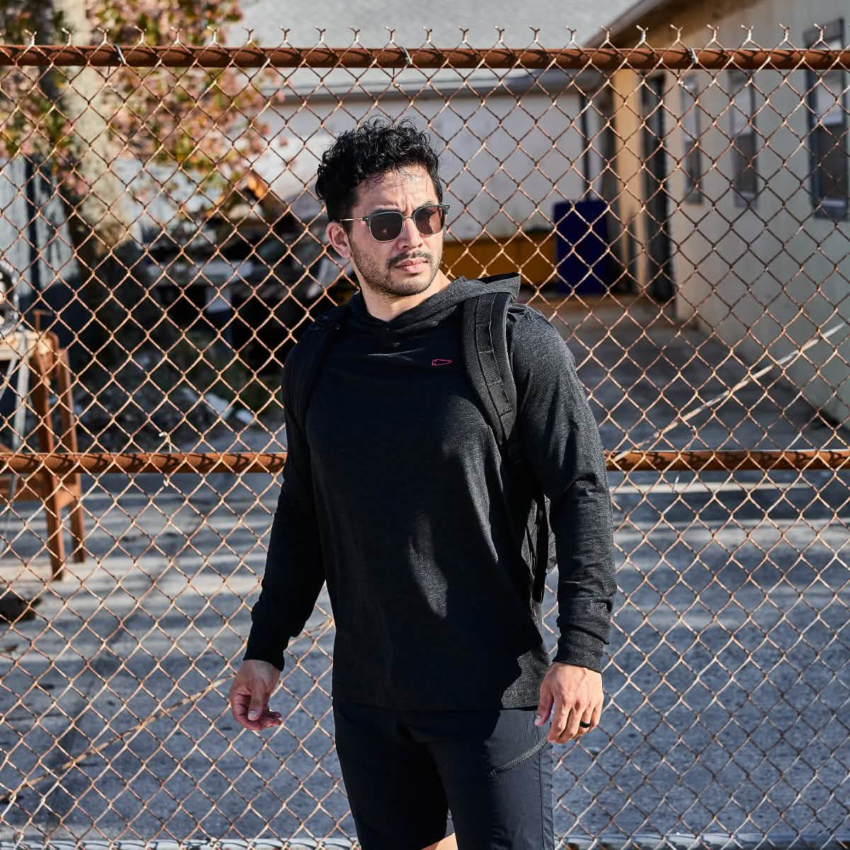 Dressed in a sleek GORUCK Subtle Spearhead Hoodie - Tri-Blend and sunglasses, the man stands confidently by the chain-link fence, his tri-blend backpack casually slung over one shoulder.