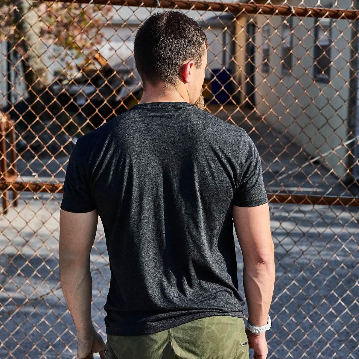 Man wearing dark gray t-shirt and camouflage pants standing in front of chain-link fence in urban setting