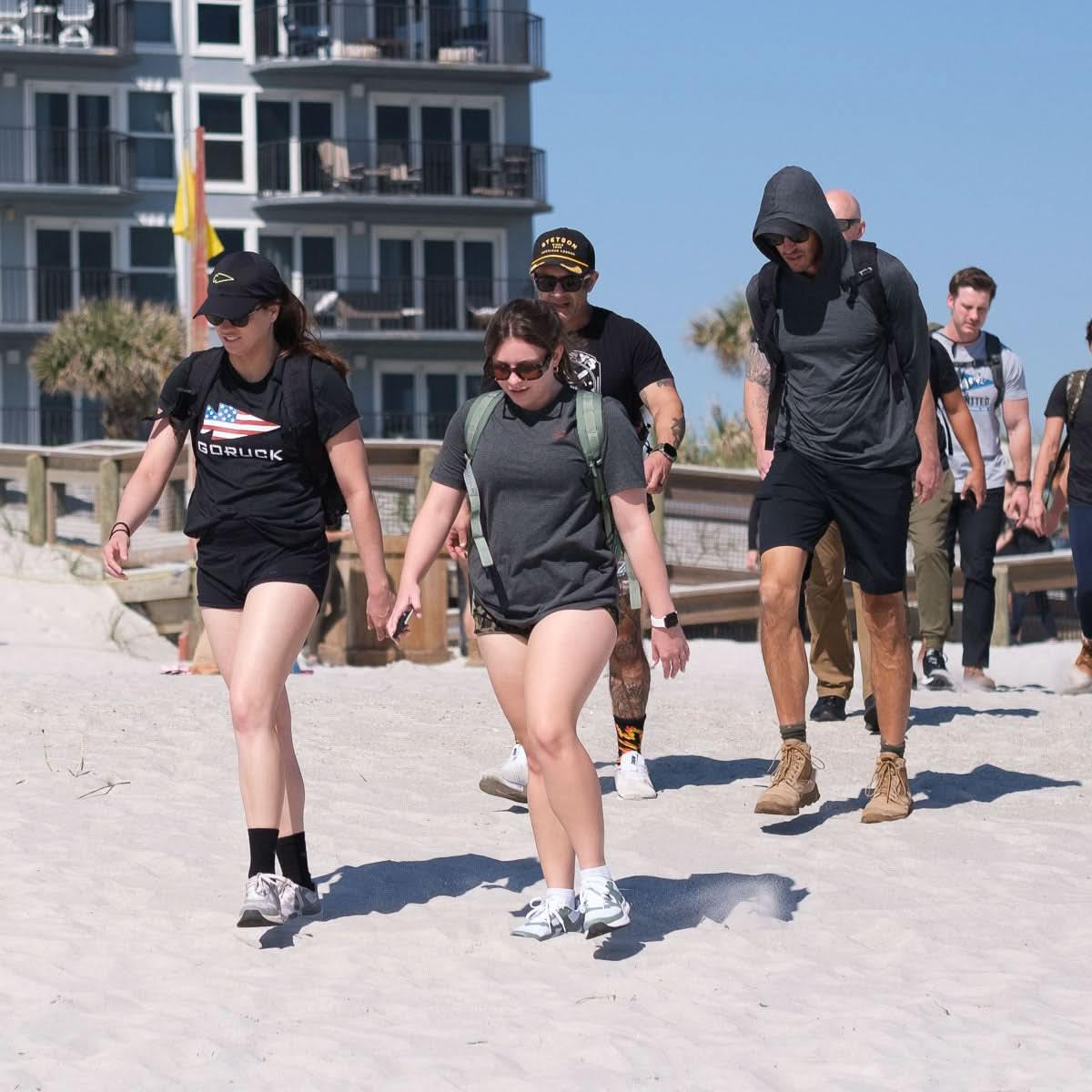 Group of diverse people hiking on sandy beach wearing casual outdoor gear and GORUCK apparel with backpacks