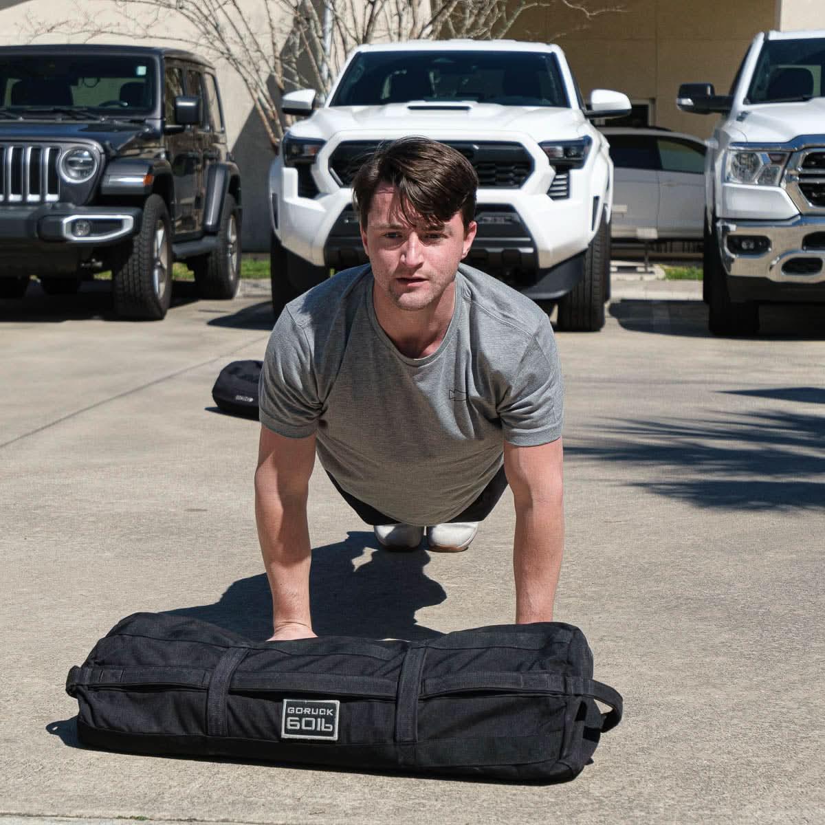 Wearing the Subtle Spearhead Tee - Unisex Tri-Blend in gray, a man does push-ups on pavement near parked cars, with a GORUCK spearhead weighted bag beside him.