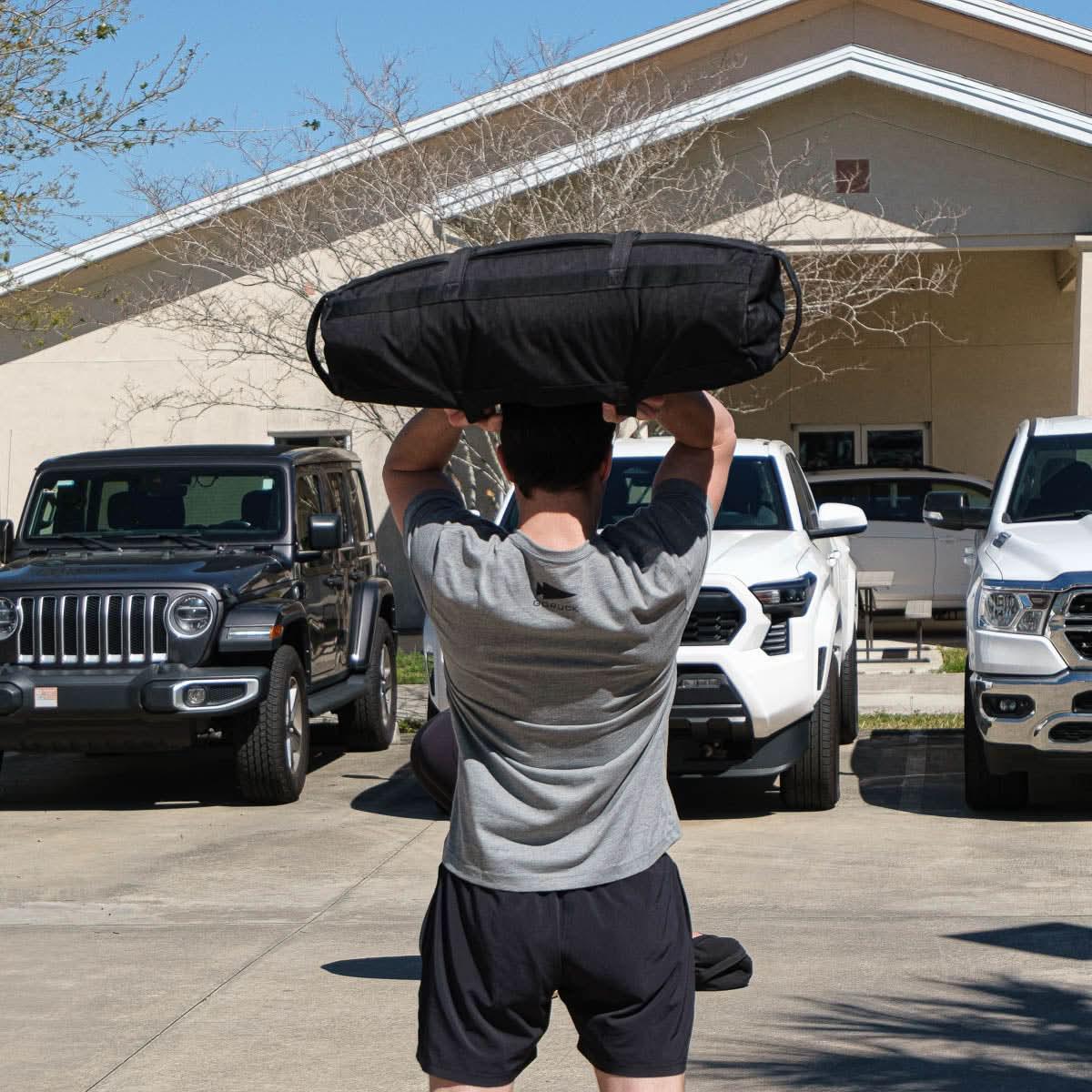 Person in gray GORUCK shirt lifting black rucksack outdoors in parking lot with vehicles and building