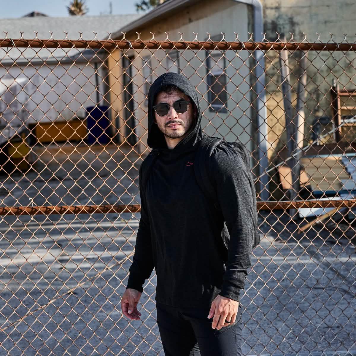 Man wearing black GORUCK hoodie with hood up and sunglasses standing in front of chain link fence outdoors
