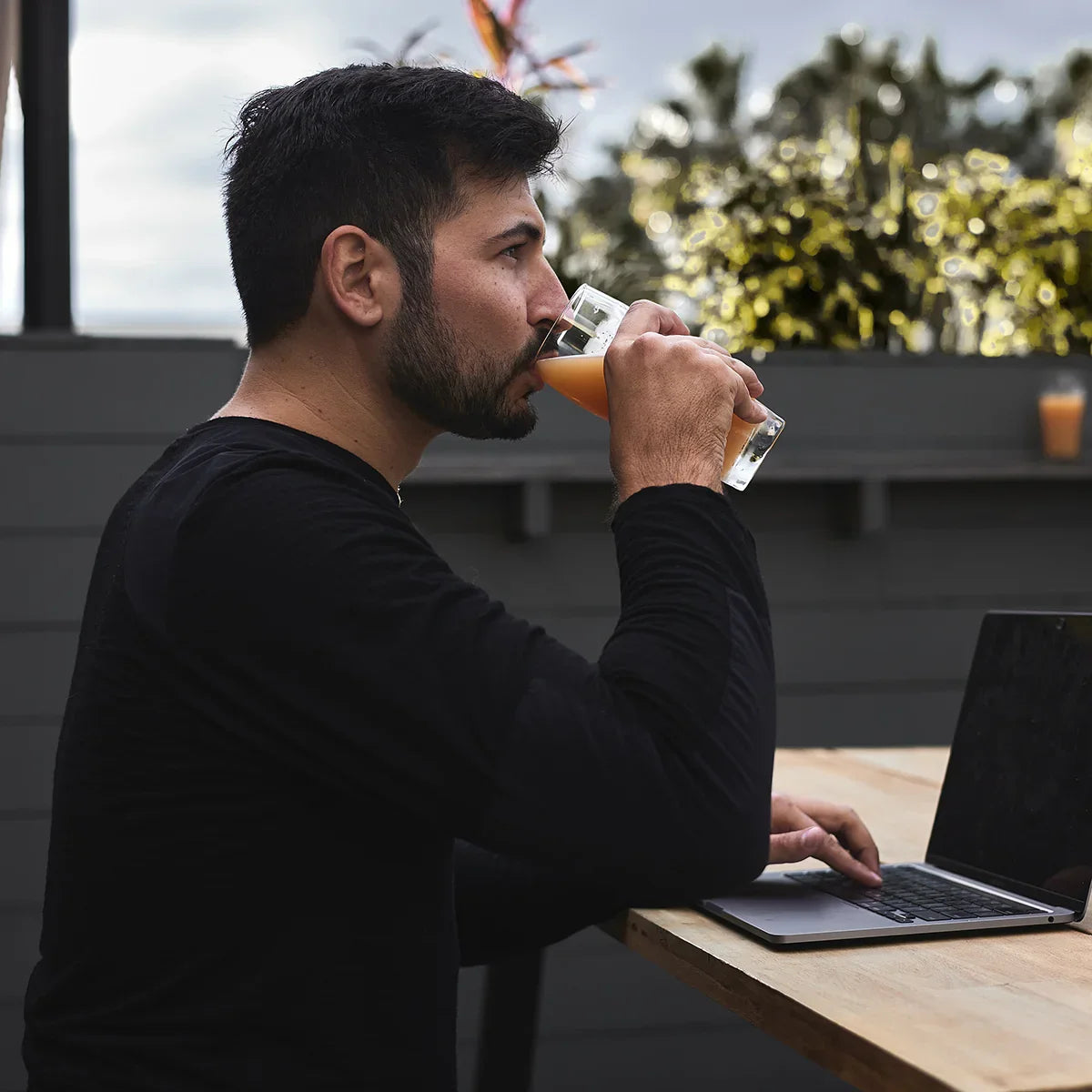 Man in black shirt drinking orange juice while working on laptop outdoors with plants background