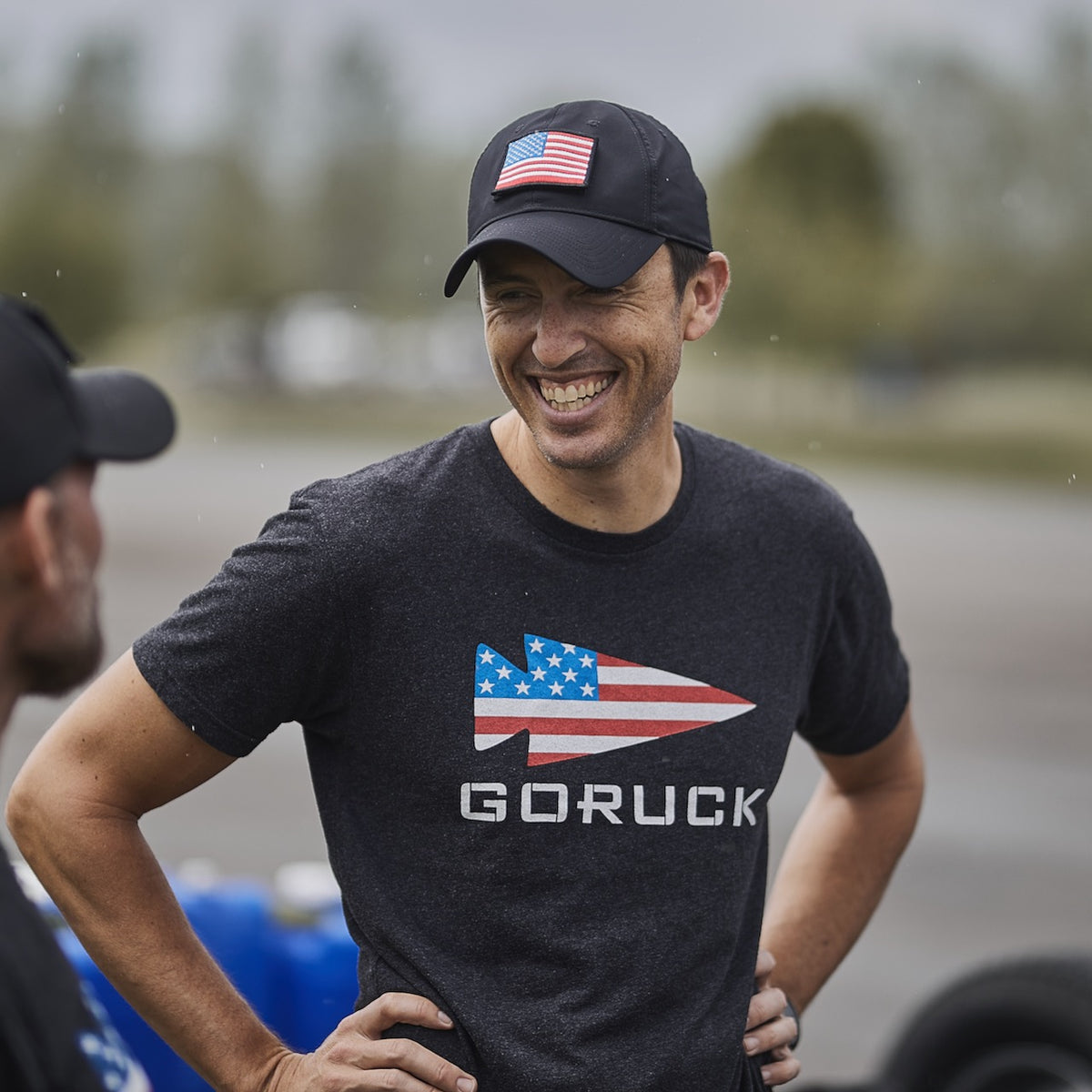 A man wearing a GORUCK shirt and the Performance TAC Hat - TOUGHDRY, styled with a USA flag, stands outdoors smiling with his hands on his hips, talking to another person.