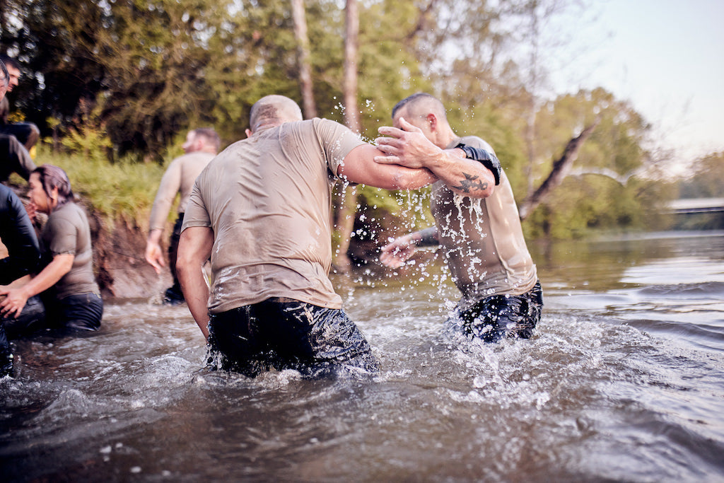 Two people grapple in shallow water, splashing as others—perhaps participants in the Team Assessment endurance event—stand nearby on the riverbank.