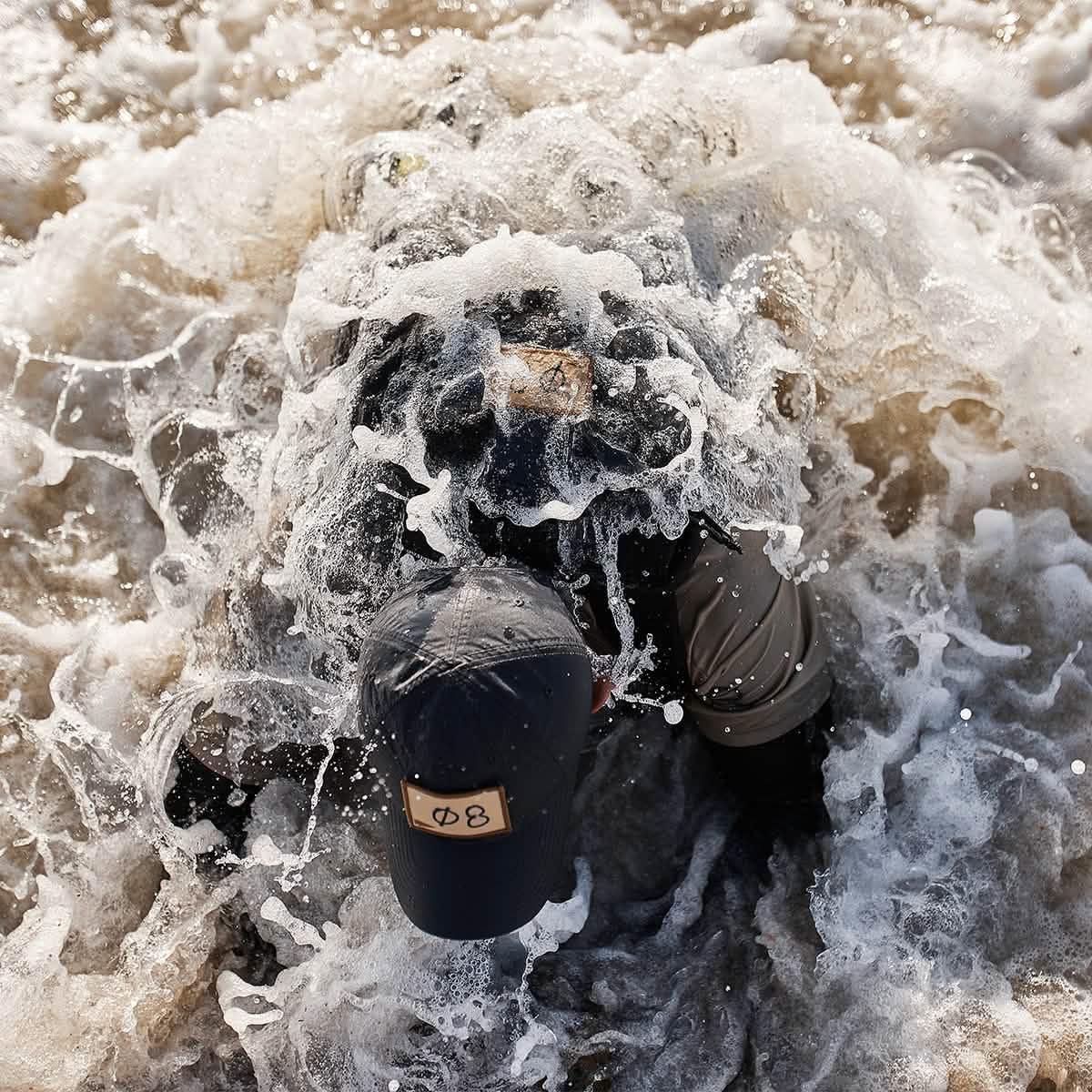 Person wearing GORUCK cap and gear submerged in frothy water during intense rucking challenge