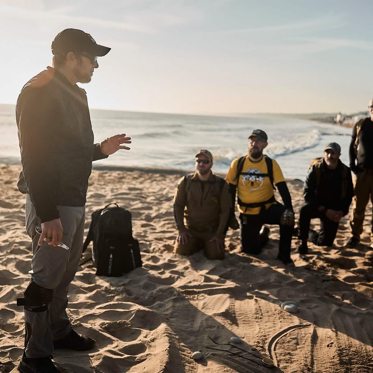 Man leading rucking training on sandy beach with group wearing tactical backpacks and outdoor gear