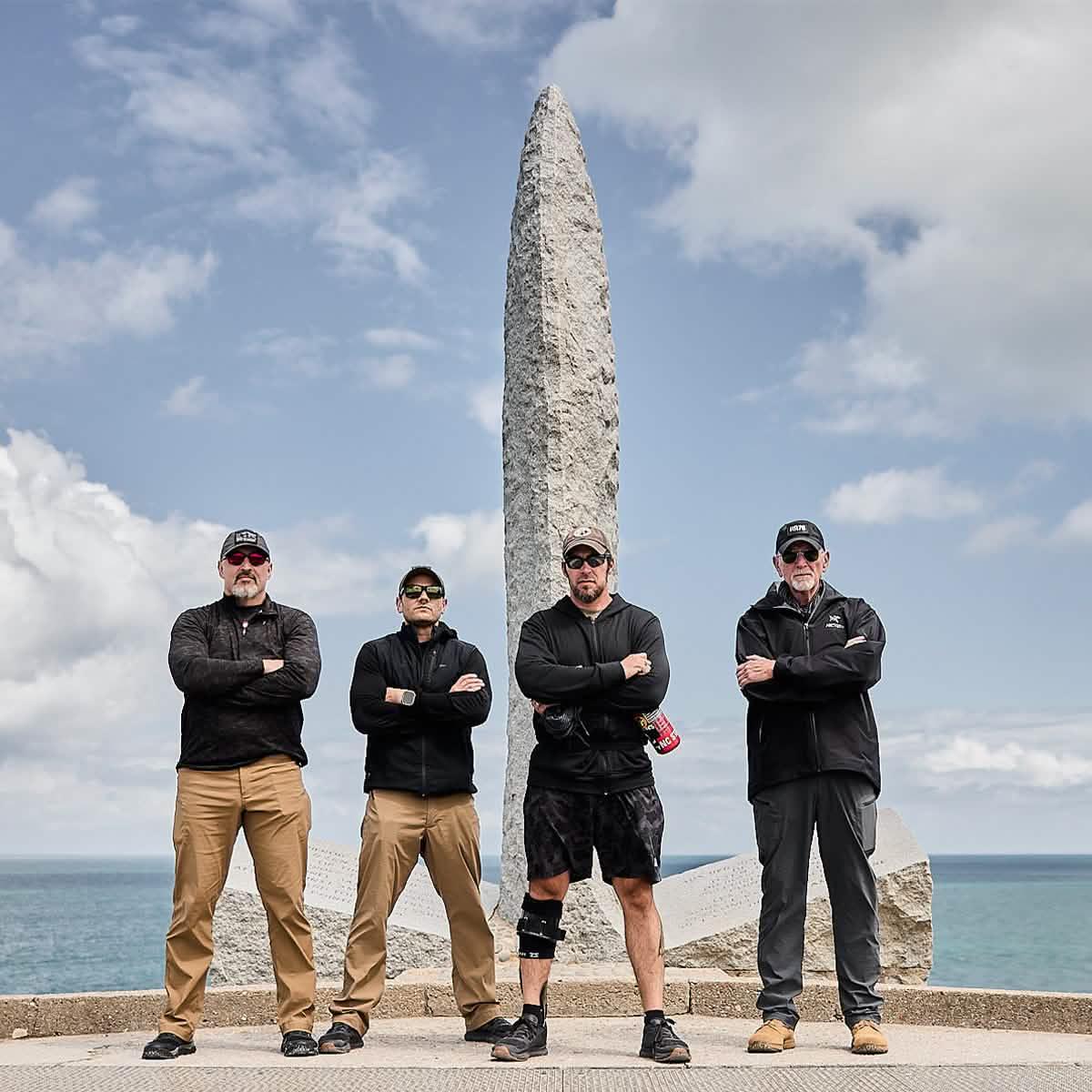 Four men wearing tactical outdoor clothing and sunglasses standing with arms crossed in front of a tall stone monument by the ocean under a cloudy sky