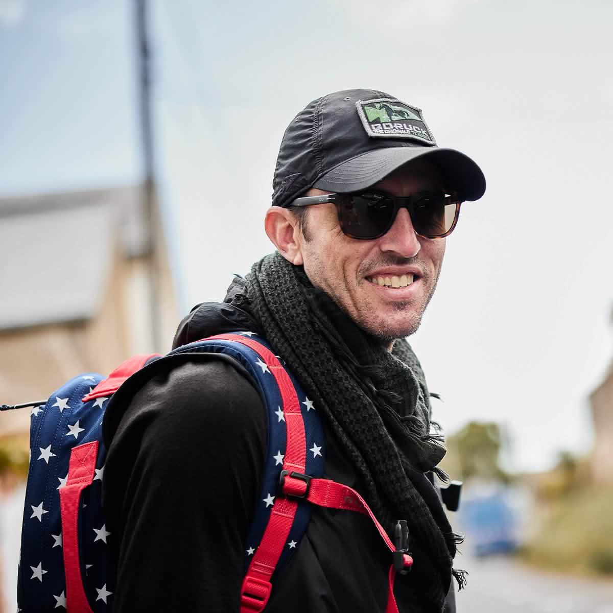 Man wearing black GORUCK cap, sunglasses, black scarf, and star-patterned blue and red GORUCK backpack outdoors