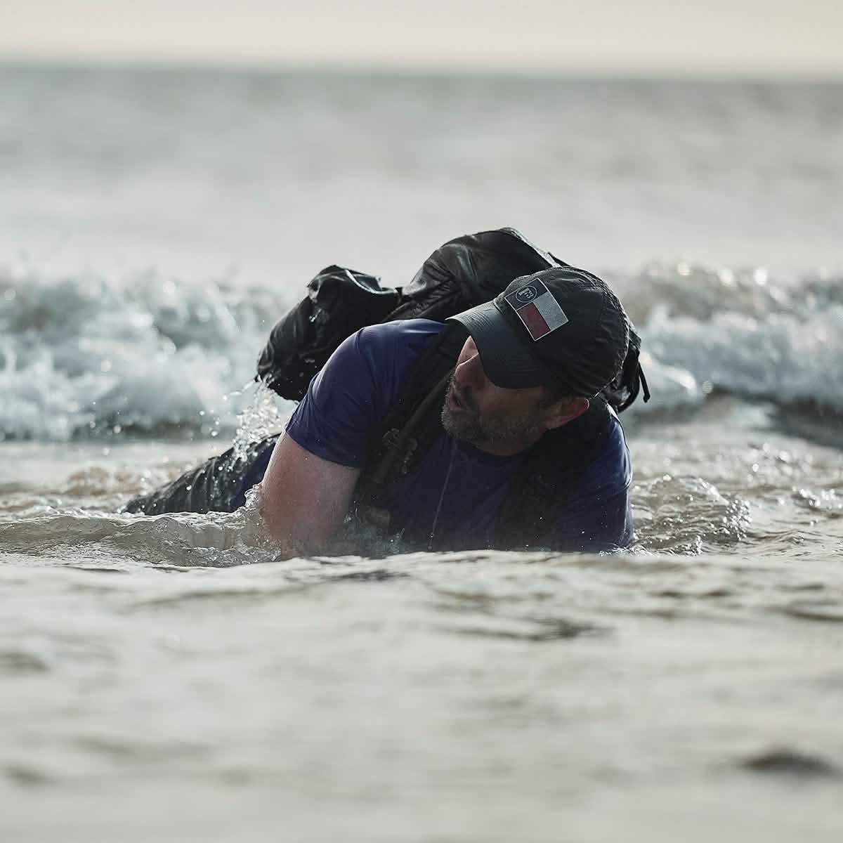 Man wearing cap and tactical backpack crawling through shallow ocean water at GORUCK military training event