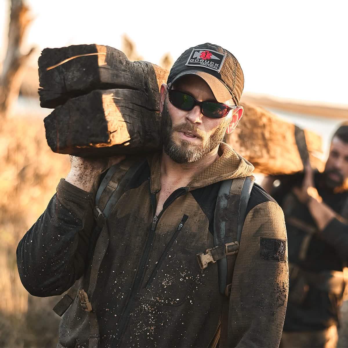 Man wearing GORUCK Tactical cap and sunglasses carrying heavy wooden logs during outdoor rucking training