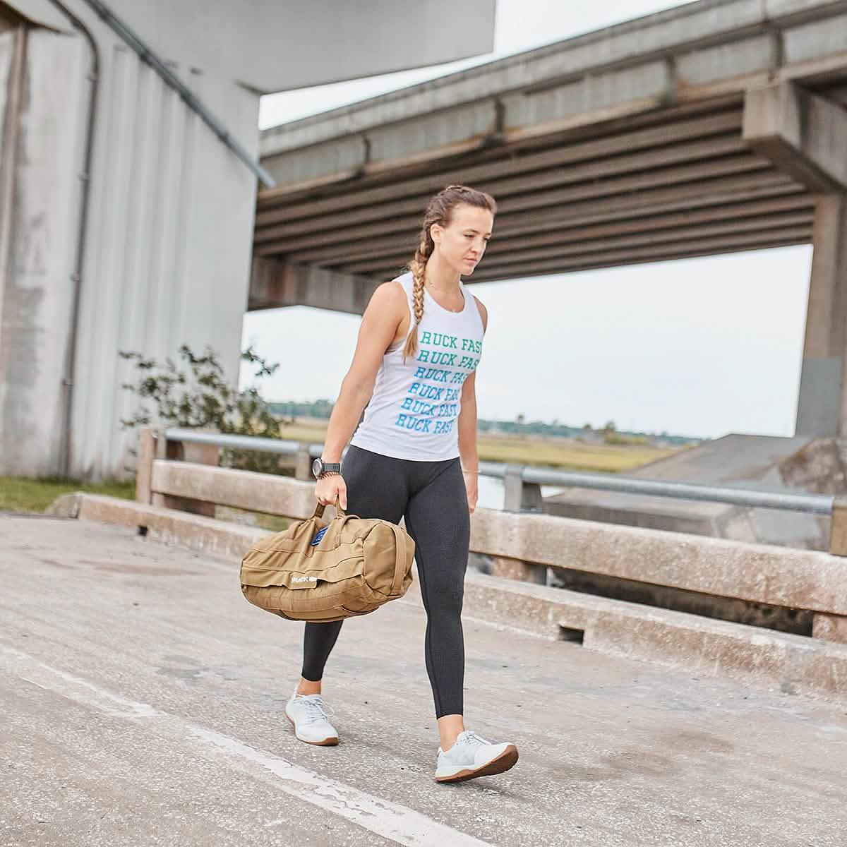 Woman carrying tan GORUCK bag walking under bridge in outdoor fitness training scene