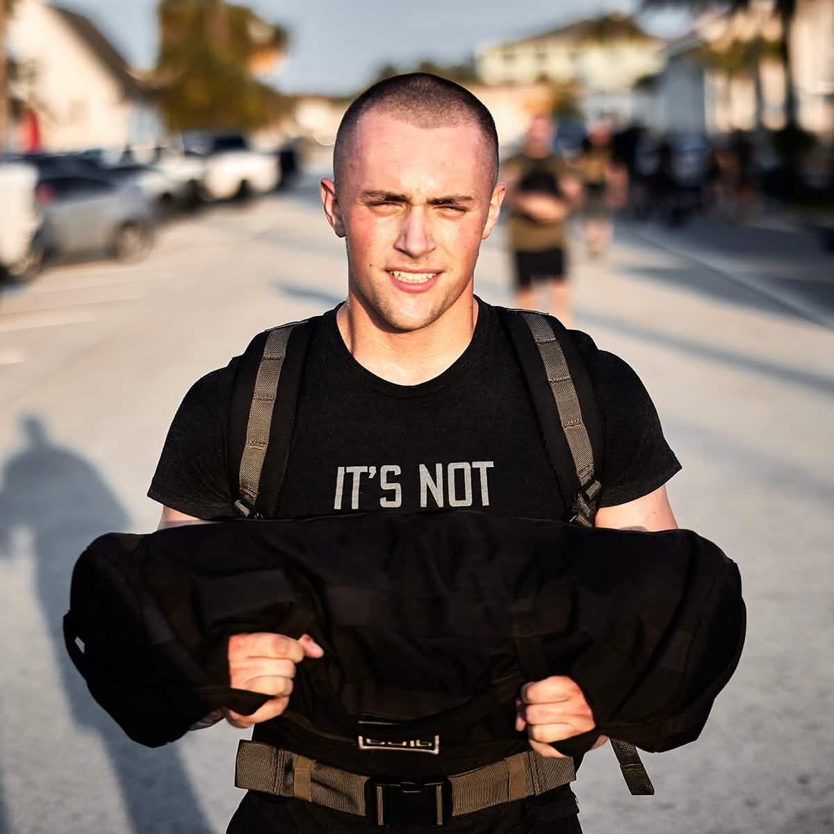 Young man with buzz cut carrying black weighted rucking bag outdoors, wearing black shirt and backpack straps