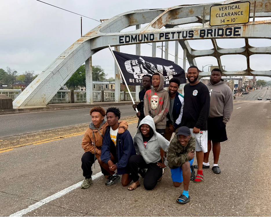 Eight young people pose and smile on the Edmund Pettus Bridge, holding a flag with text—celebrating camaraderie after participating in the Ruck Workout and Film Screening for The Give Team, inspired by GORUCK HQ in Jacksonville Beach, FL.