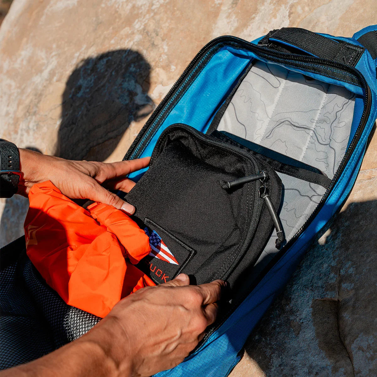 Hands packing a black GORUCK pouch and an orange item into a blue backpack with a topographic map interior
