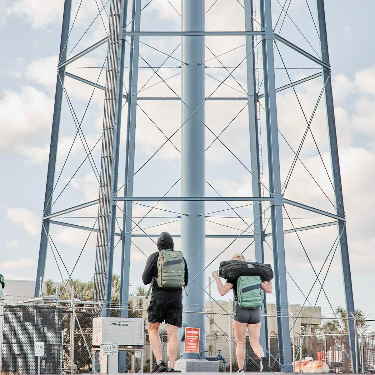 Two people rucking with GORUCK backpacks and gear under a large outdoor metal structure