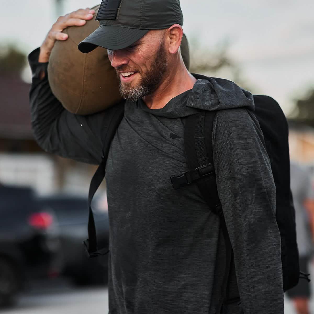 Man wearing GORUCK ToughMesh charcoal hoodie, cap, and backpack carrying a sandbag outdoors