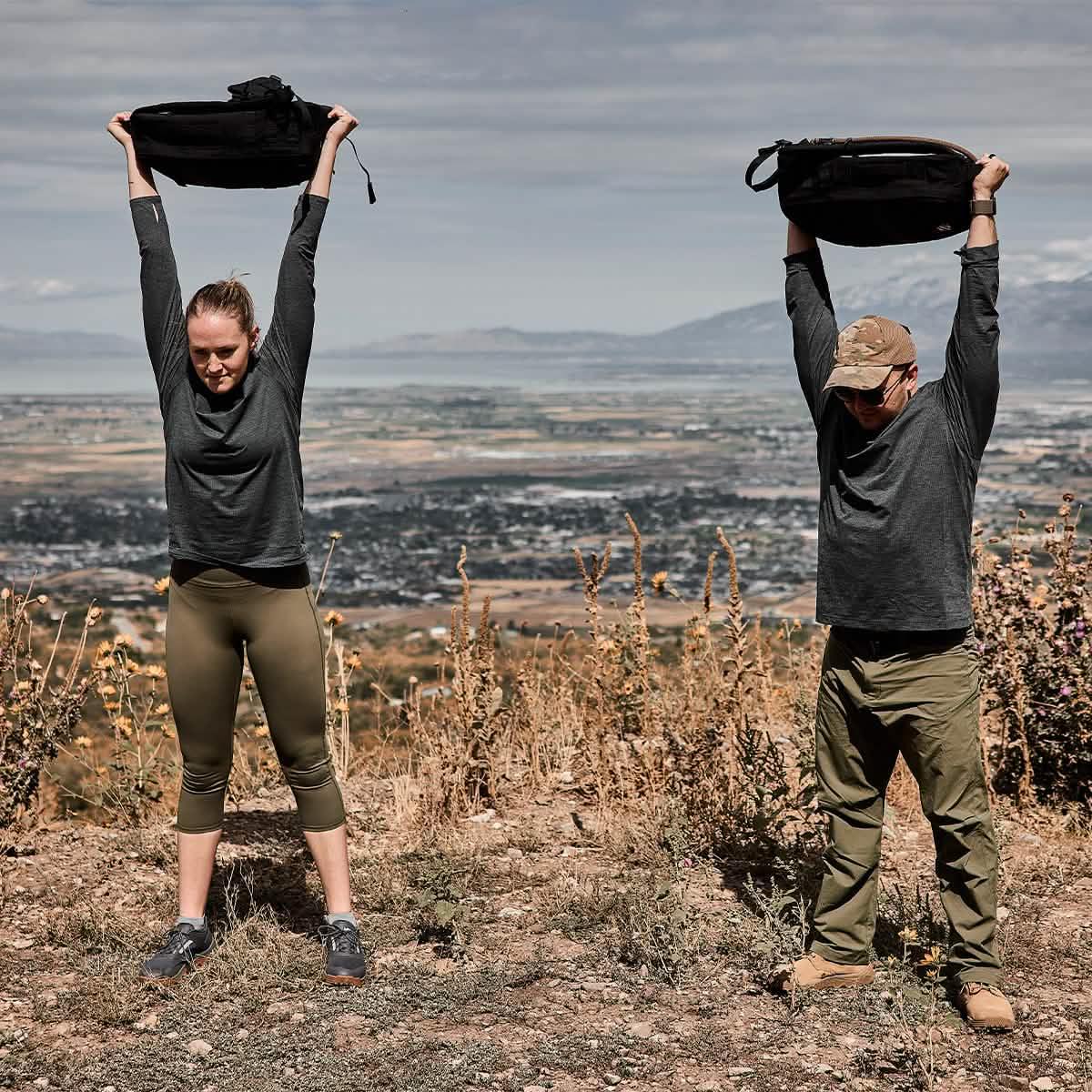 Two people in GORUCK ToughMesh long sleeve shirts rucking outdoors on a dirt trail, lifting backpacks overhead, with a scenic valley and mountains in the background.