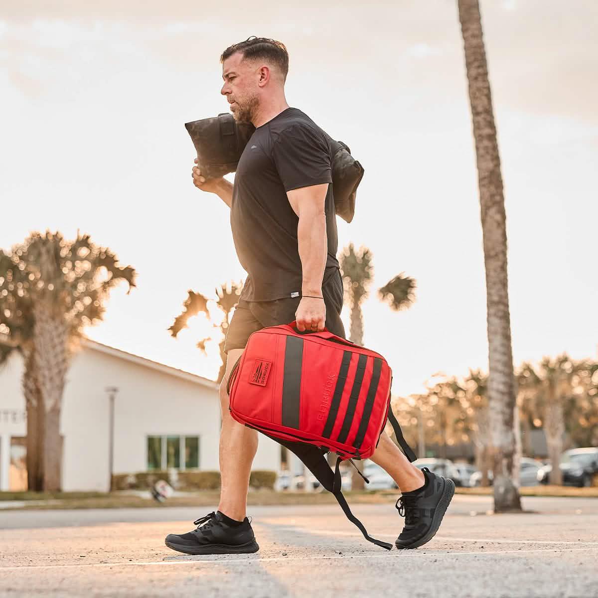 Man walking outdoors carrying a red GORUCK rucksack and a black weighted sandbag at sunset