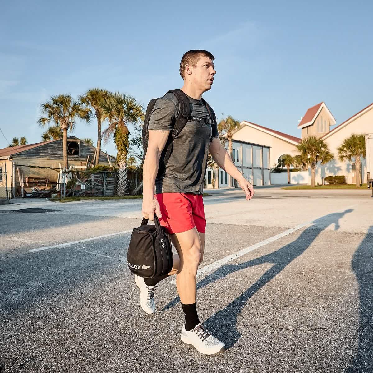 Man walking outdoors carrying a black 35 lb GORUCK tough mesh sandbag with palm trees and buildings in background