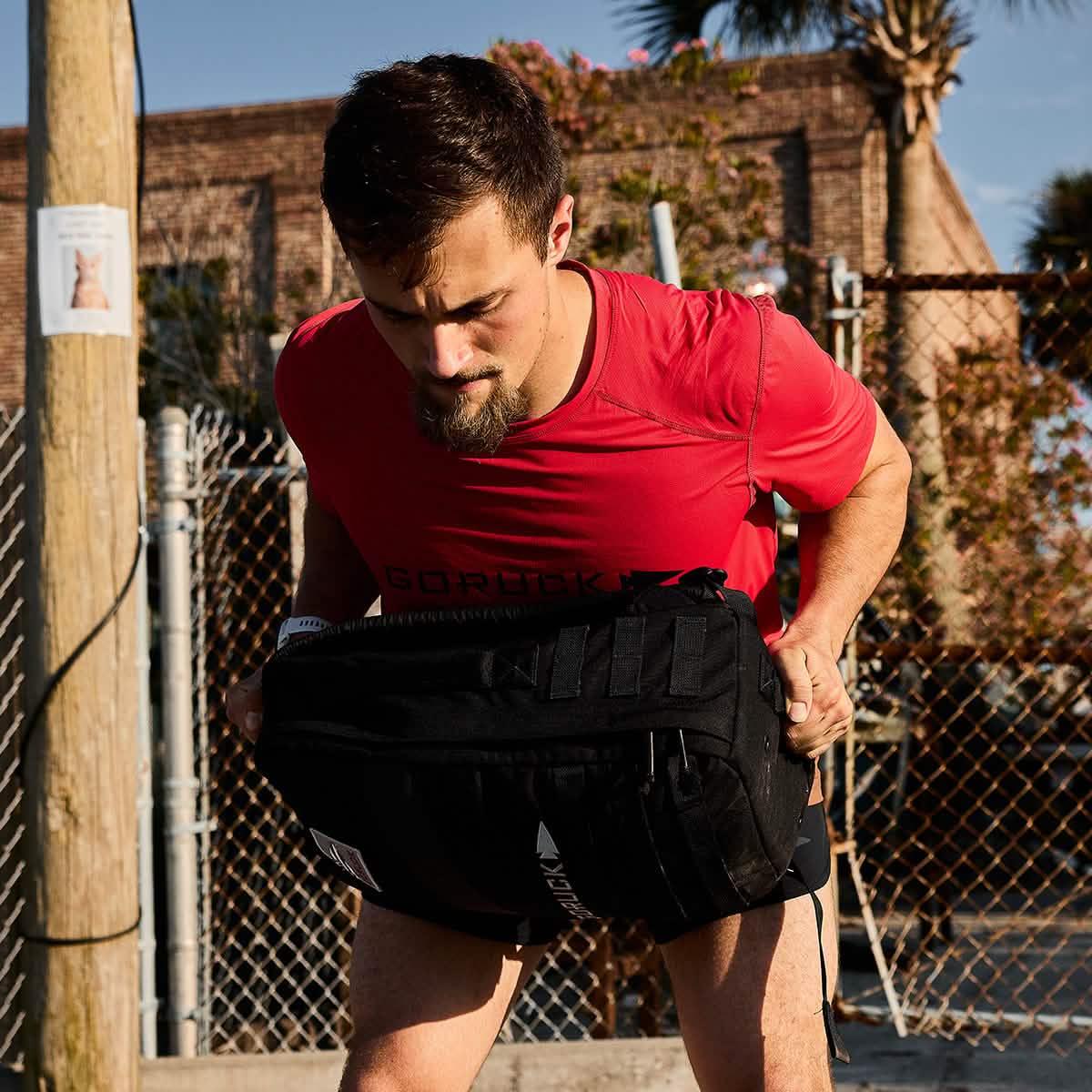 Man in red GORUCK shirt lifting a black weighted rucksack outdoors near chain link fence