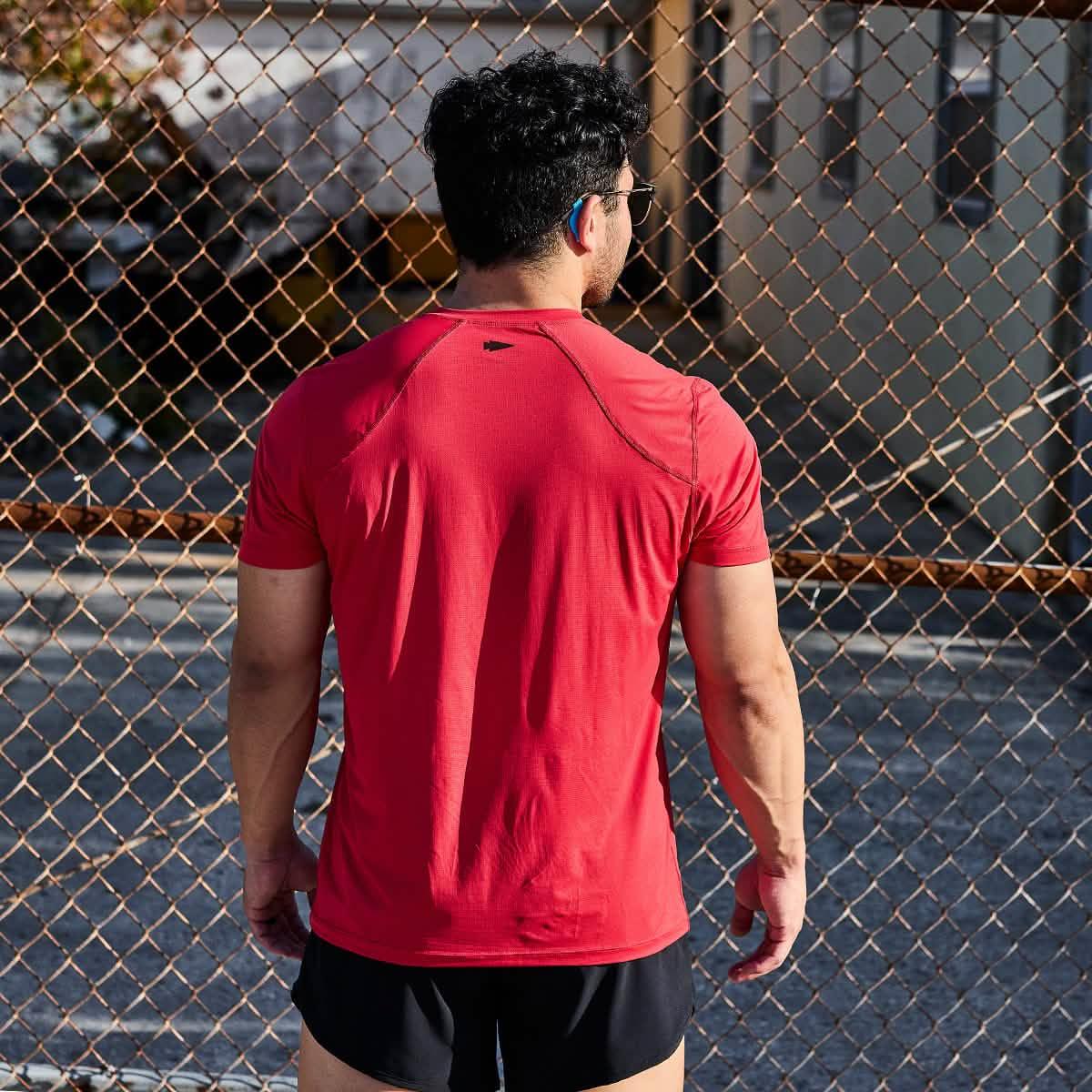 Man wearing red athletic shirt and black shorts stands facing chain-link fence in outdoor urban setting