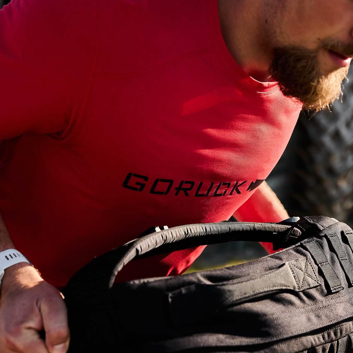 Close-up of a man wearing a red GORUCK shirt holding a black rucking bag during outdoor training