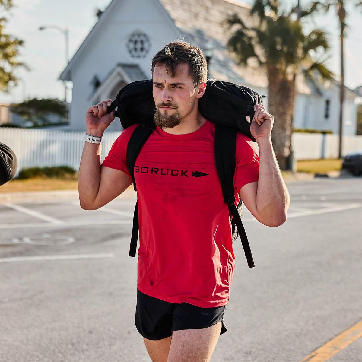 Man wearing red GORUCK t-shirt and black shorts carrying black rucking sandbag on shoulders outdoors