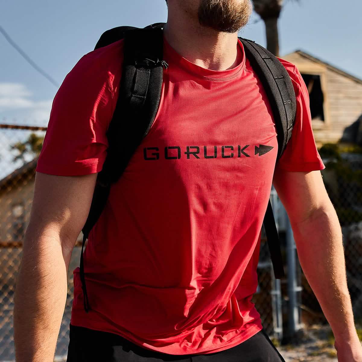Close-up of a person wearing a red GORUCK t-shirt and black backpack outdoors near a chain-link fence and building