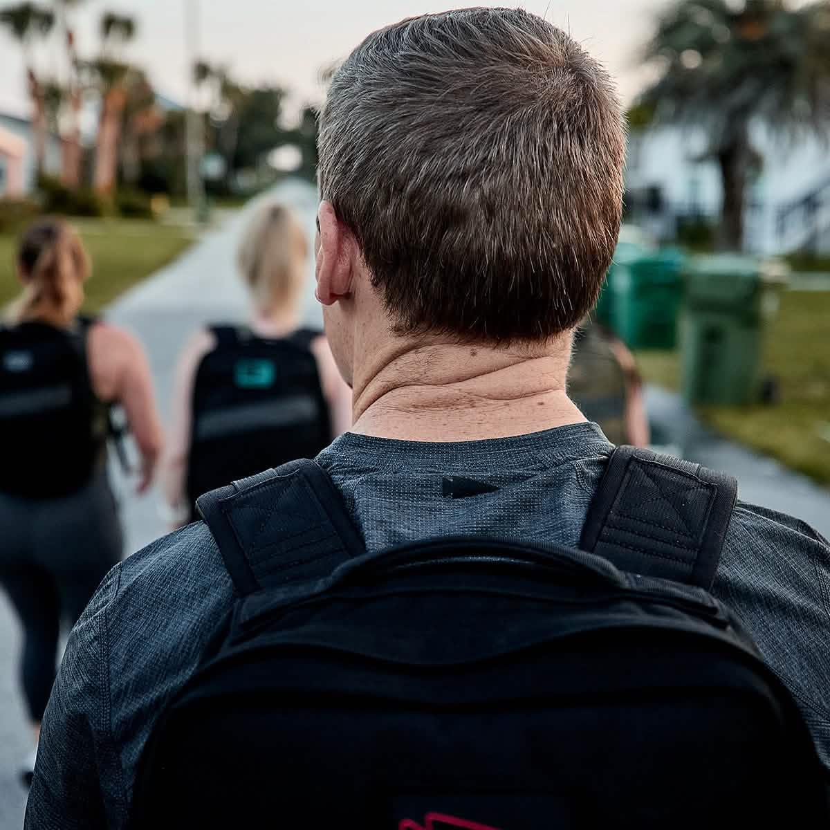 Back view of man wearing GORUCK backpack with two women walking ahead on a suburban sidewalk