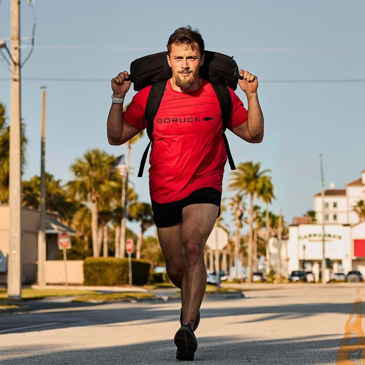 Man running on street with GORUCK red shirt and black weighted rucking bag during sunny day