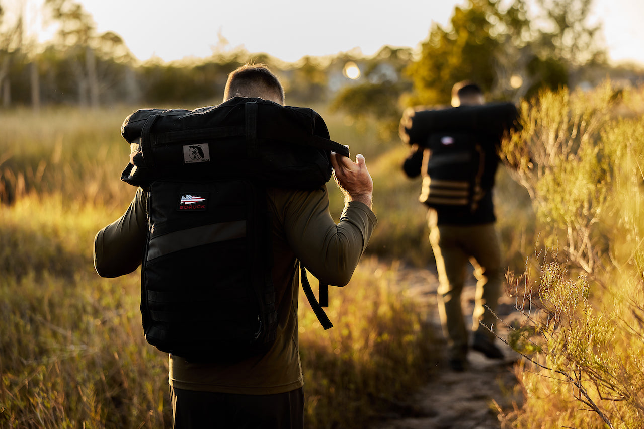 Two people hike on a narrow trail through tall grass, each carrying a large black backpack on their shoulders.