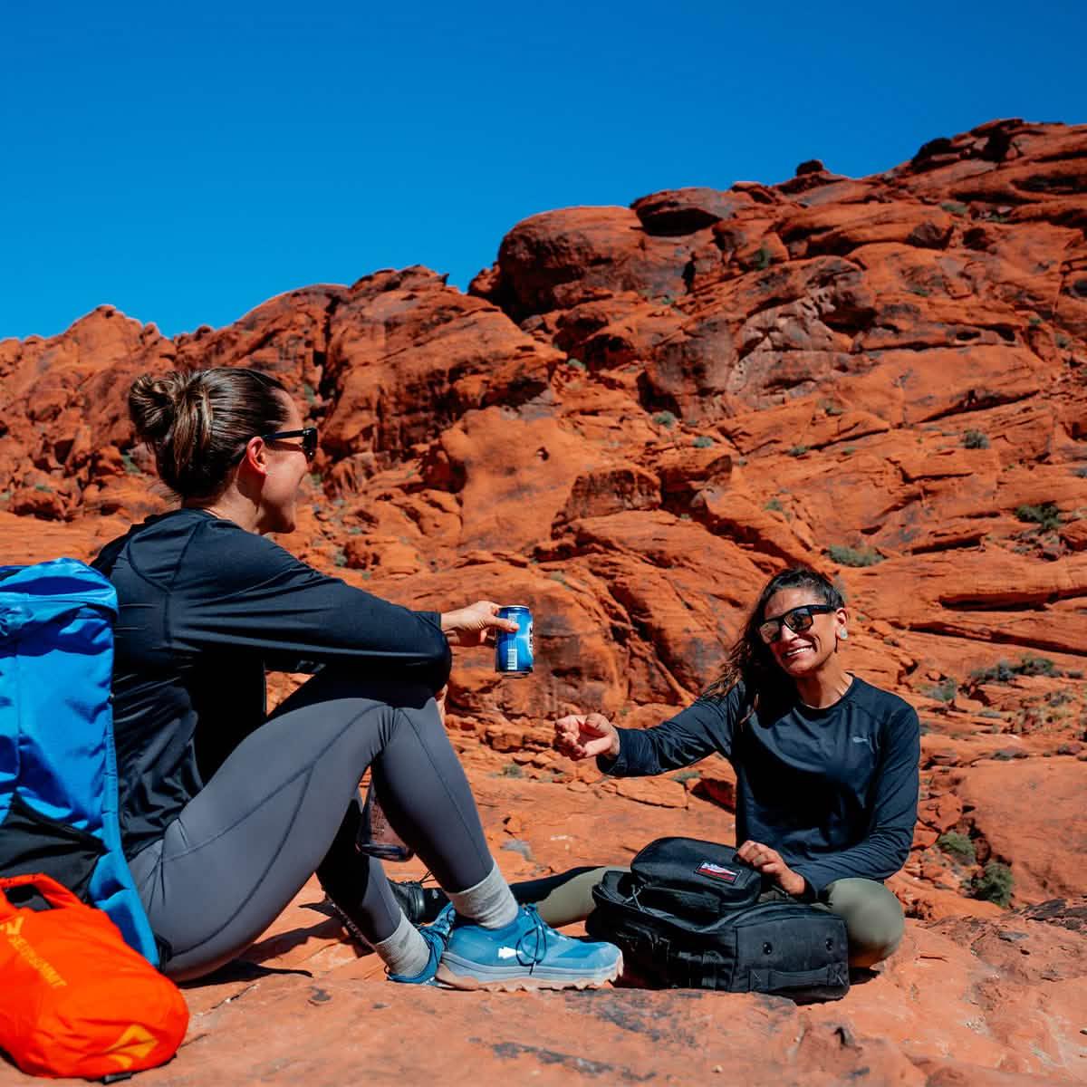 Two women with GORUCK packs relaxing on red rocks, enjoying a hiking break outdoors