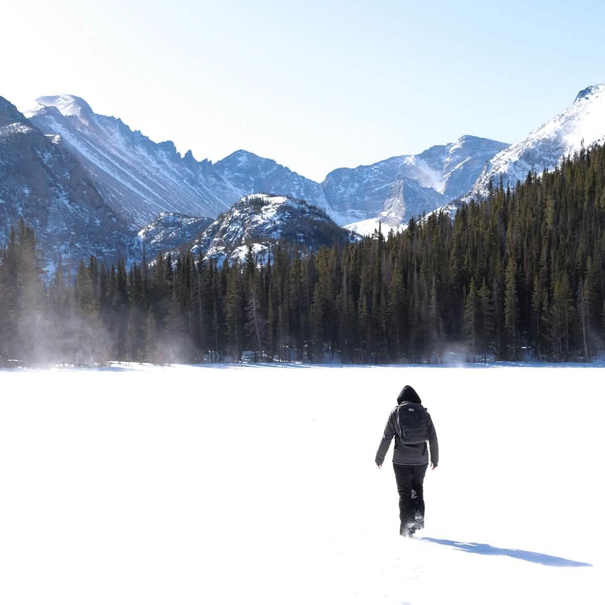 Person hiking in snowy winter landscape with backpack, pine forest, and snow-covered mountains