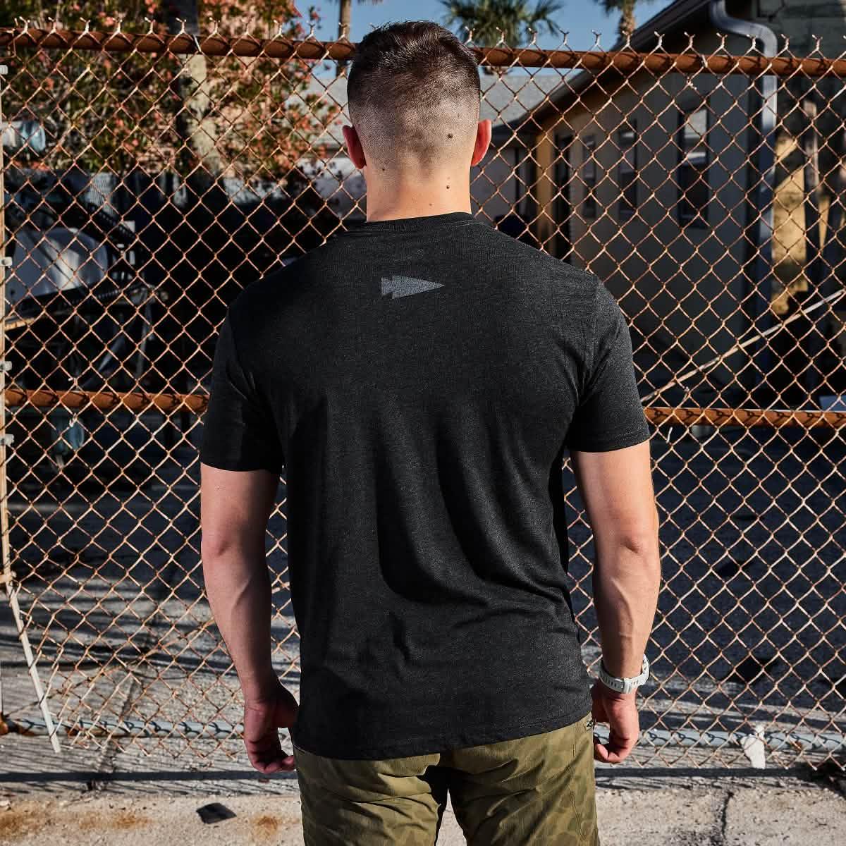 Man wearing black GORUCK t-shirt and olive camo pants standing facing chain link fence outdoors