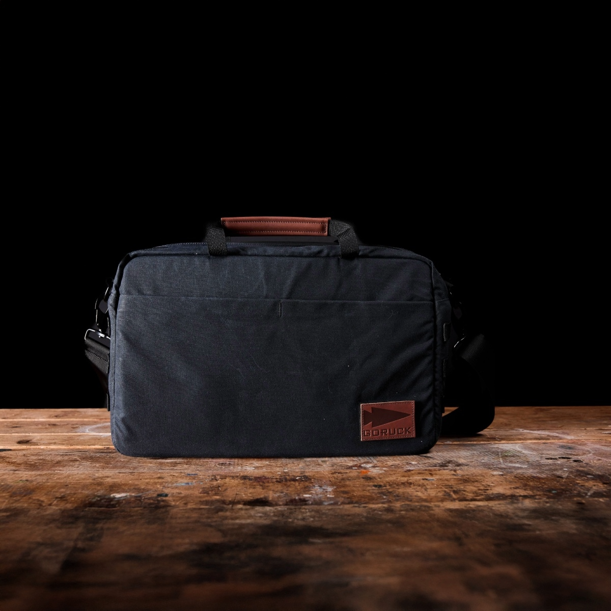 The Shoulder Bag - Waxed Canvas features a black design with a brown leather handle, crafted for asymmetrical carries. It sits on a rustic wooden surface against a black background, showcasing its rugged and stylish appeal.