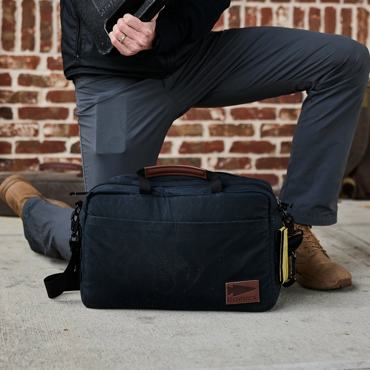 A person kneels on one knee behind a black Shoulder Bag - Waxed Canvas placed on the ground in front of a brick wall, ready for asymmetrical carries.
