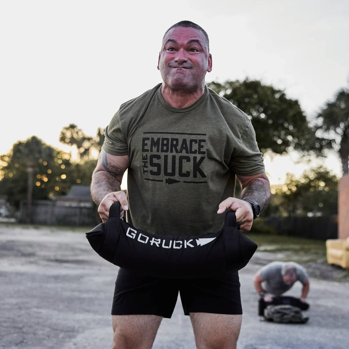 Man wearing an olive GORUCK t-shirt with Embrace The Suck text lifting a weighted GORUCK sandbag during outdoor fitness training