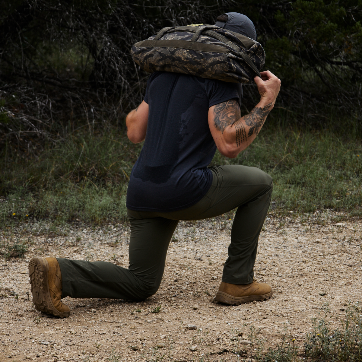 A man kneels outdoors with a camo-patterned sandbag on his shoulders, wearing boots and Men’s Simple Pants - Lightweight ToughDry® as part of his athletic gear.