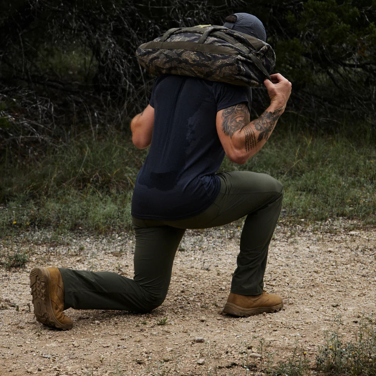 Person in black shirt and green pants doing lunges outdoors carrying a camo GORUCK sandbag on shoulders