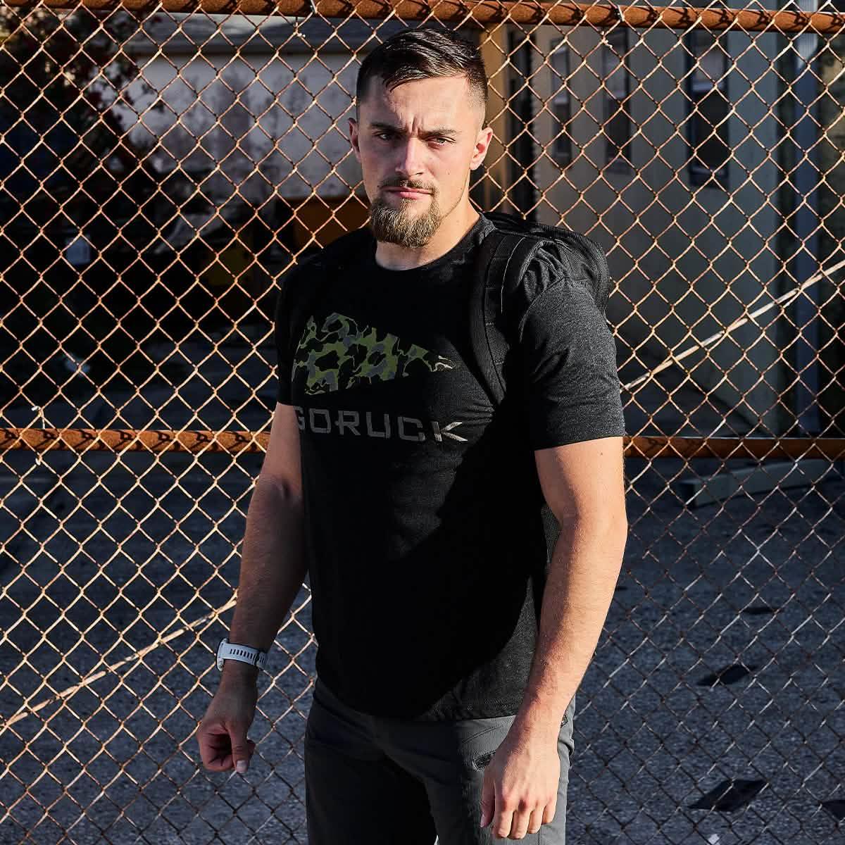 Athletic man wearing black GORUCK t-shirt with green frogskin camo logo and backpack, standing by chain-link fence