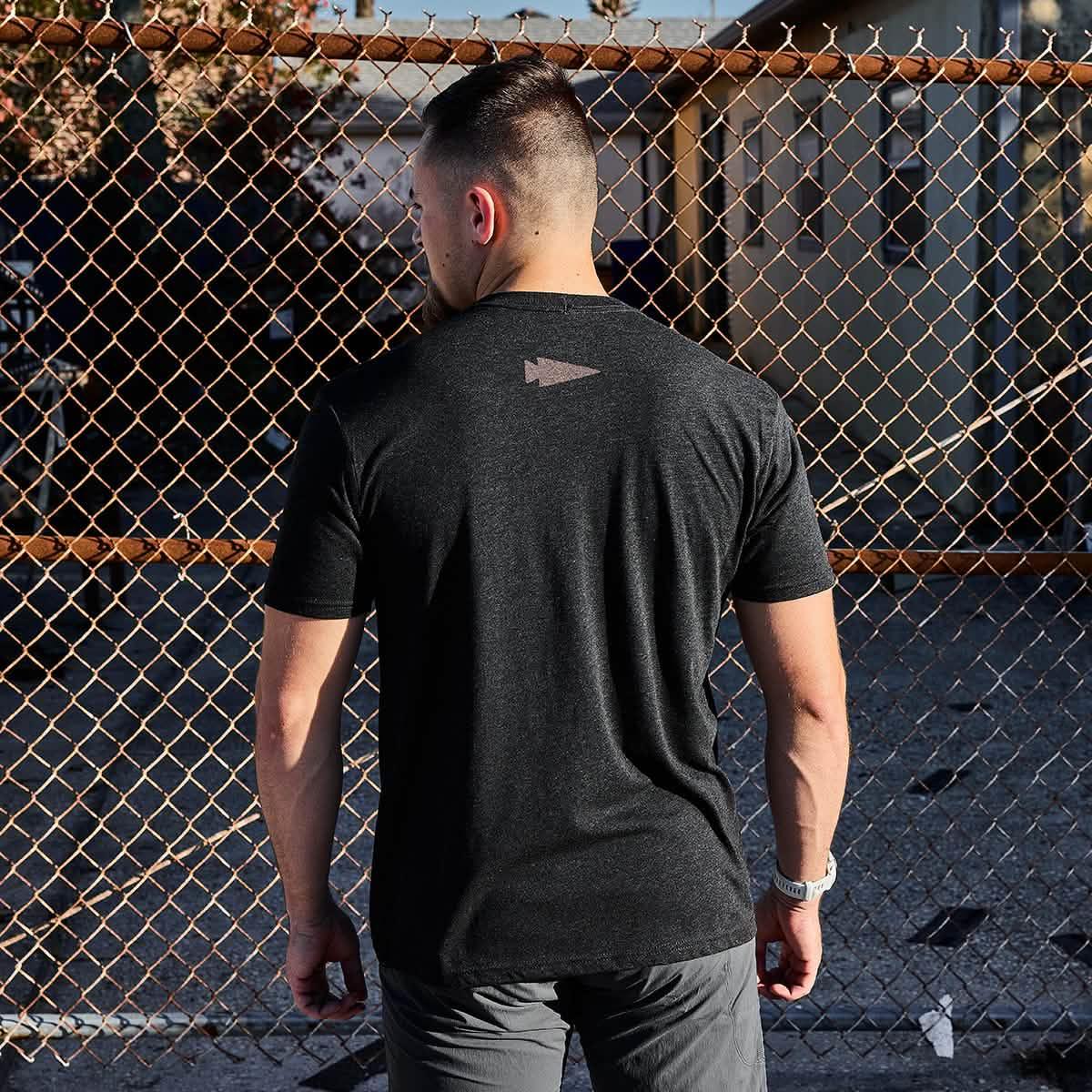 Man wearing black GORUCK t-shirt with logo on back standing in front of rusty chain link fence in outdoor urban setting